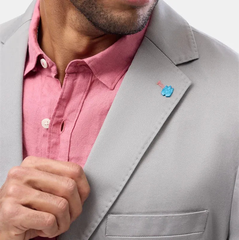 Close-up of a man wearing a light gray suit jacket over a pink button-down shirt, with a small blue and red flower lapel pin on the jacket lapel.