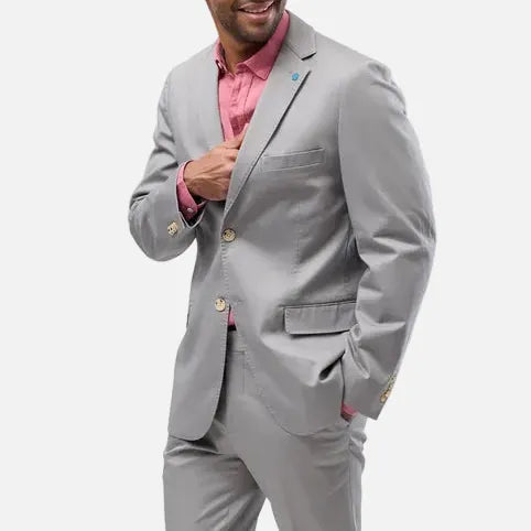 A man modeling a light gray three-piece suit with a pink shirt and brown leather shoes, standing against a solid light gray background.