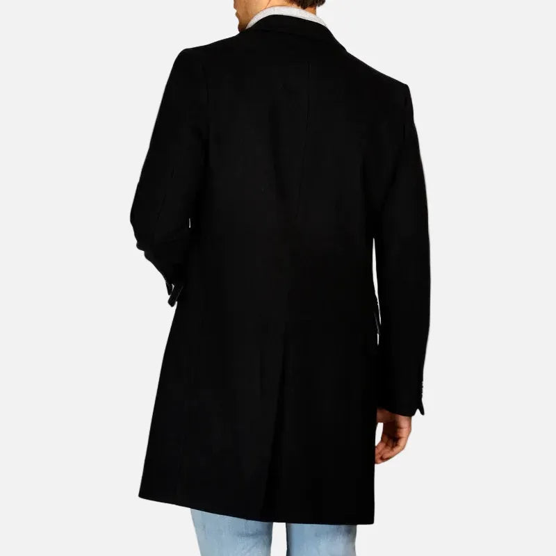 A man shown from the back wearing a black mid-length overcoat over a white collared shirt and light blue jeans, set against a plain light gray studio background.