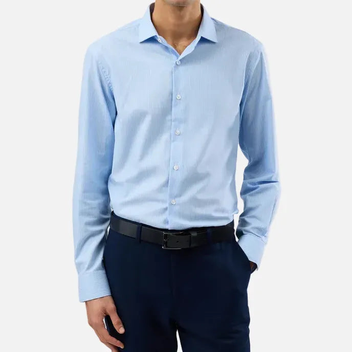 A man wearing a light blue long-sleeve dress shirt, dark navy trousers, and a black belt, standing against a light gray studio background.