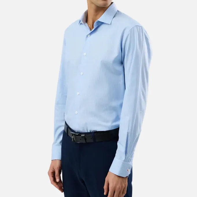 A man wearing a light blue long-sleeve dress shirt, dark navy trousers, and a black belt, standing against a plain light gray studio background.