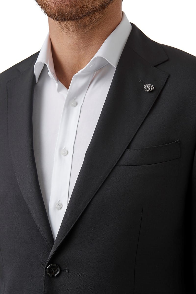 A close-up of a man wearing a black suit jacket over a white dress shirt, with a small circular lapel pin visible on the jacket.