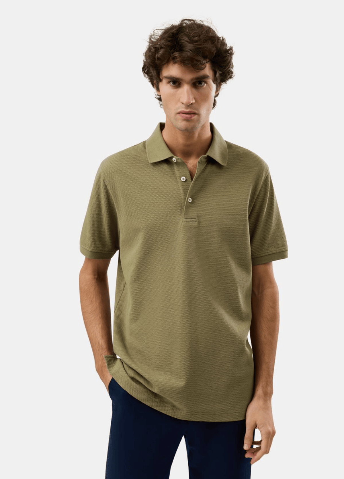 A man wearing a short-sleeve polo shirt in olive green, paired with dark navy pants, standing against a light gray studio background.