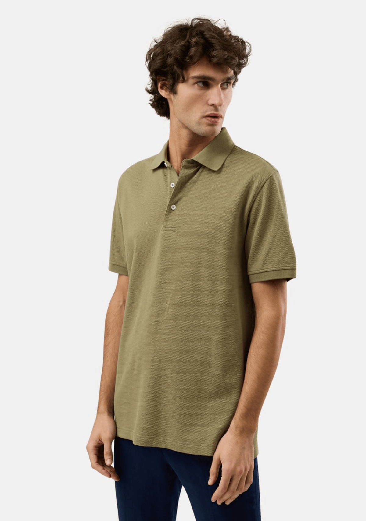 A man wearing a short-sleeve polo shirt in olive green, paired with dark blue pants, photographed against a clean light gray studio background.