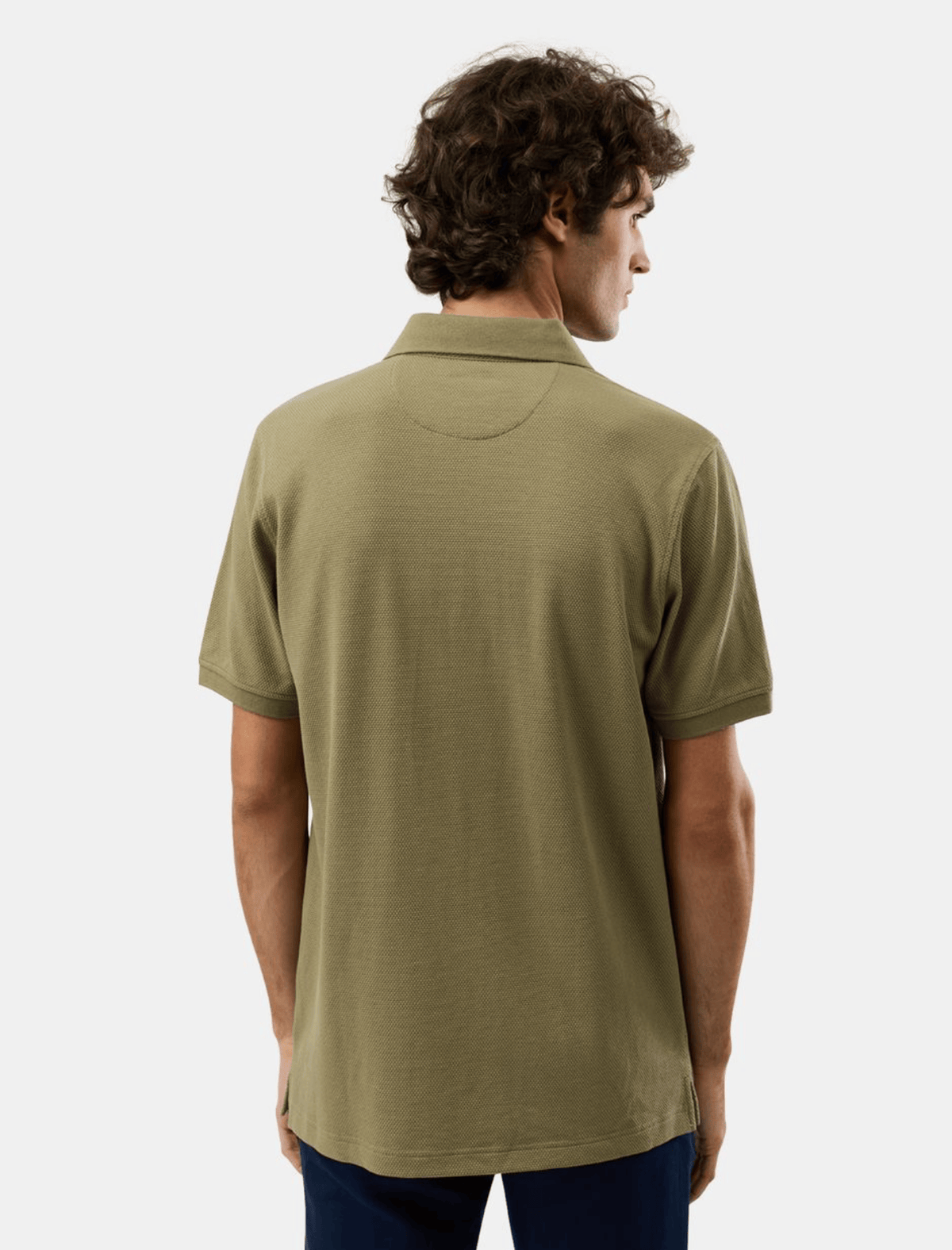 A man viewed from the back wearing a short-sleeve polo shirt in olive green, paired with dark blue pants, against a light gray studio background.