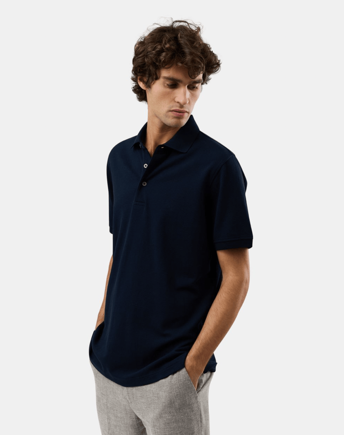 A man wearing a dark navy blue short-sleeve polo shirt and light gray trousers, standing against a plain light gray studio background.