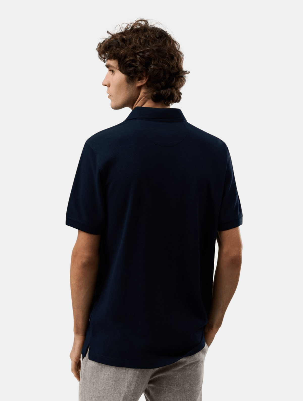 A man with curly brown hair is shown from the back wearing a dark navy blue short-sleeve polo shirt and light gray trousers, set against a plain light gray studio background.