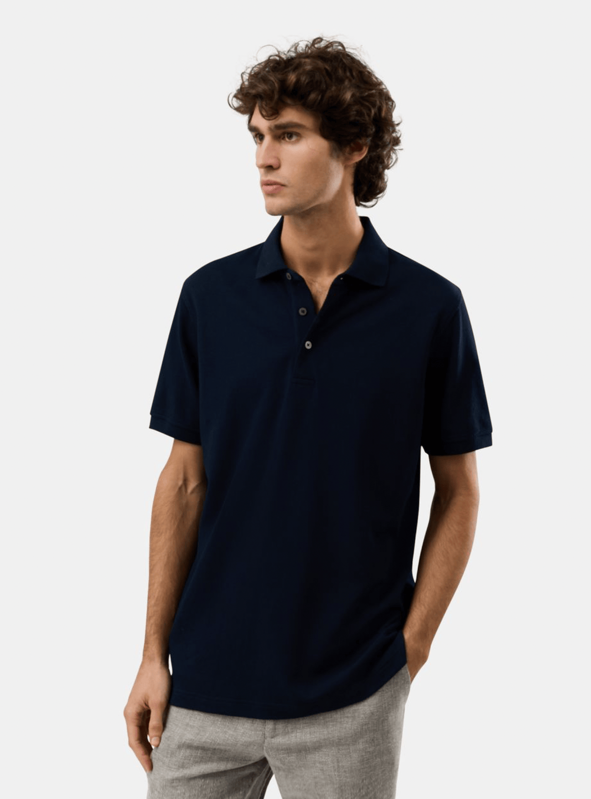 A man wearing a dark navy blue short-sleeve polo shirt and light gray trousers, standing against a plain light gray studio background.