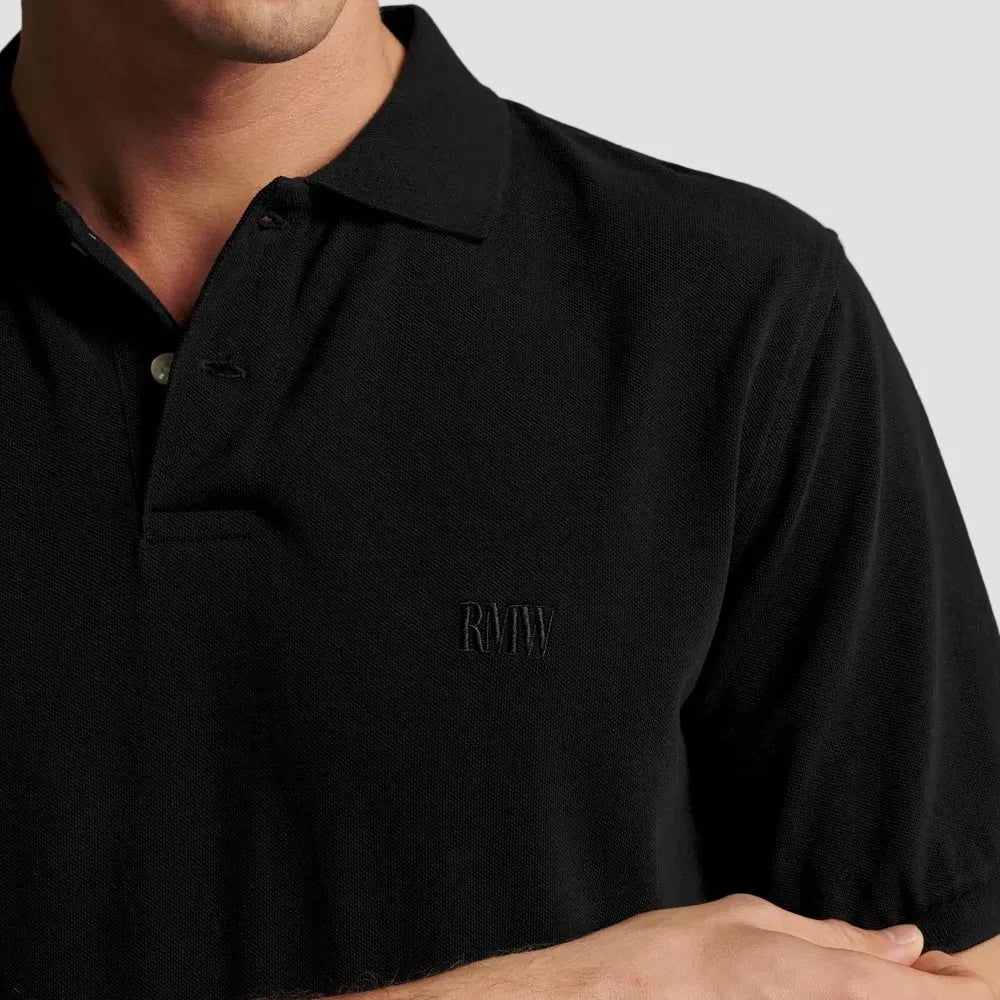 Close-up of a man wearing a black short-sleeve polo shirt with a subtle tonal embroidered logo on the left chest, set against a plain beige background.