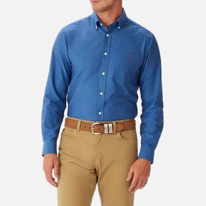 A man wearing a blue long-sleeve button-down shirt, khaki pants, and a brown belt, standing against a plain white background.