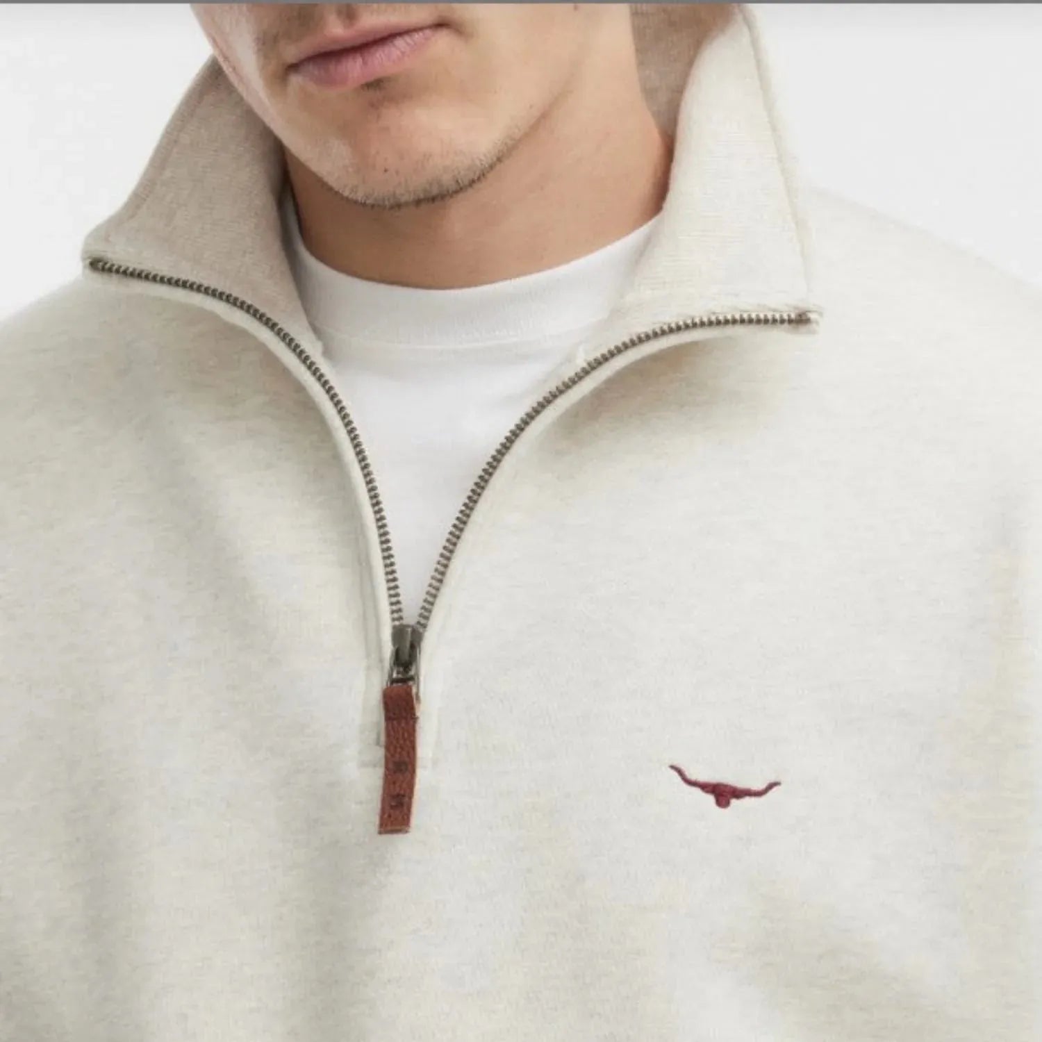 Close-up of a man wearing a cream-colored quarter-zip sweatshirt with a brown leather zipper pull and a small embroidered longhorn logo on the chest.