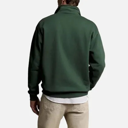 A man viewed from behind wearing a dark green quarter-zip fleece jacket, beige trousers, and brown shoes, standing against a solid beige studio background.