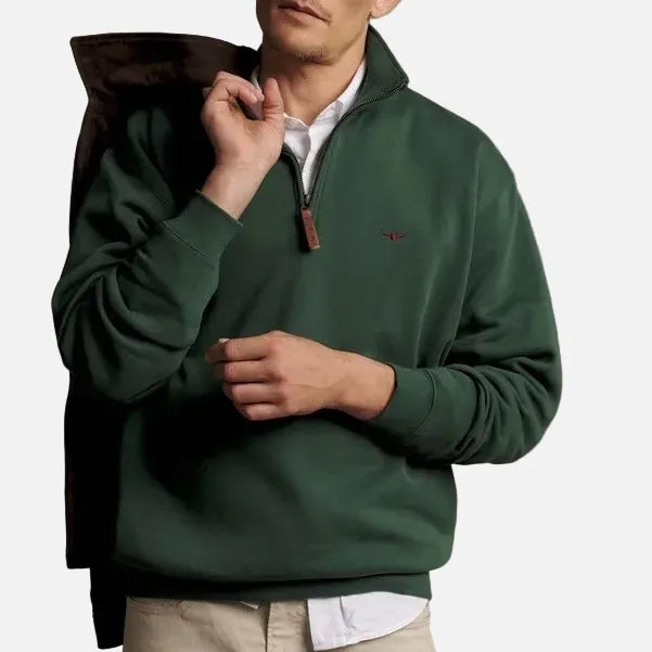 A man wearing a dark green quarter-zip pullover sweater over a white collared shirt, with a dark brown jacket draped over his shoulder, set against a plain beige studio background.