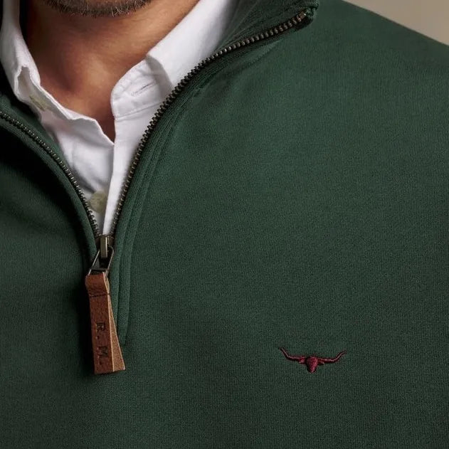 Close-up of a man wearing a dark green quarter-zip pullover sweater over a white collared shirt. The sweater has a brown leather zipper pull tab and a small embroidered red bull logo on the left chest.