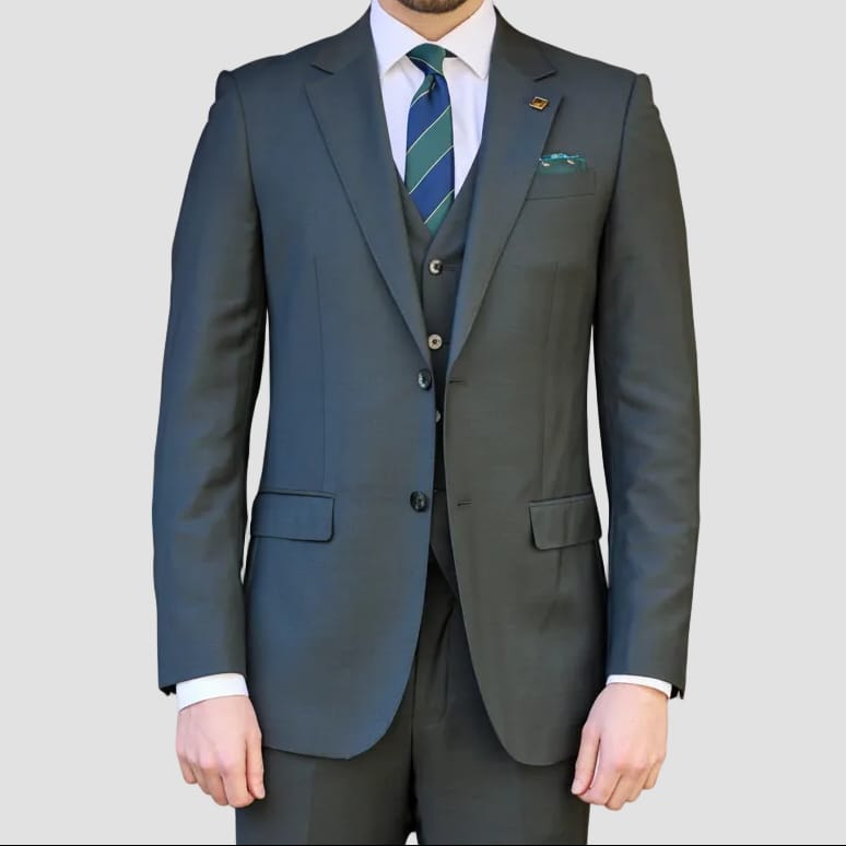 A man wearing a dark slate blue three-piece suit with a matching vest and trousers, a white dress shirt, and a green and blue striped tie, standing against a solid gray background.
