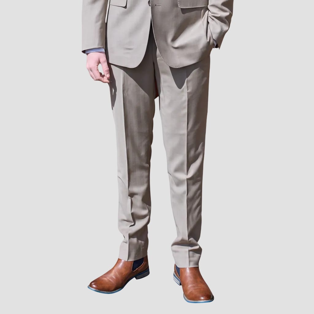A man wearing light beige suit trousers with a matching blazer and brown leather Chelsea boots, shown from the waist down against a solid light gray background.