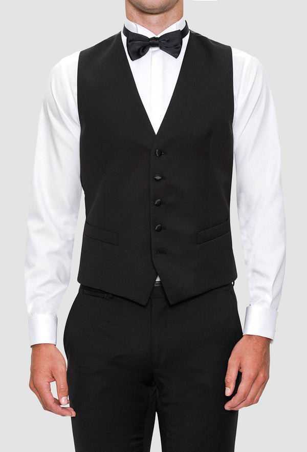 a model wears the Joe Black slim fit mail vest in black pure wool styled with a white shirt and black bow tie