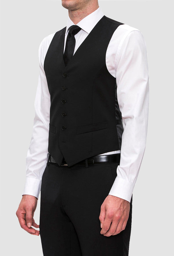 a side on view of a model wearing the Joe Black slim fit mail vest in black pure wool with a white shirt underneath and a black tie